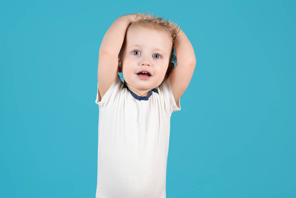 Симпатичная кудрявая двухлетняя девочка поднимает руки вверх и держит голову. Изолированный на синем фоне
 - Фото, изображение