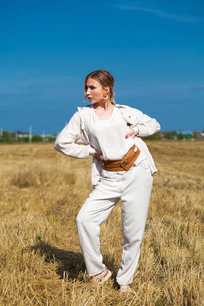 Beautiful blonde girl in a beige suit posing on a background of haystacks in a cut field. - Photo, Image