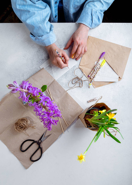 Γυναίκα συσκευάζοντας ανοιξιάτικα λουλούδια ως δώρα, γράφοντας καρτ-ποστάλ σε λευκό τραπέζι. Κάτοψη, προσανατολισμός πορτραίτου - Φωτογραφία, εικόνα