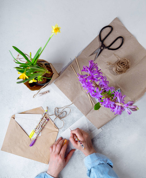 Woman in casual clothes packing spring potted flowers in brown paper as gifts, writing poscards. Top view, verical image - Photo, Image