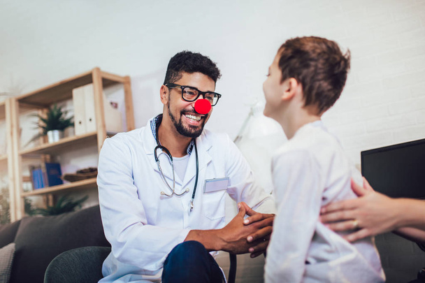 Афроамериканский педиатр со стетоскопом и клоунским носом разговаривает с маленьким мальчиком
  - Фото, изображение