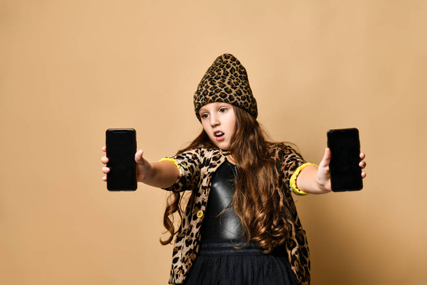Young plus size girl in leopard colored casual clothing, accessories and black skirt standing and holding two smartphones - Foto, imagen