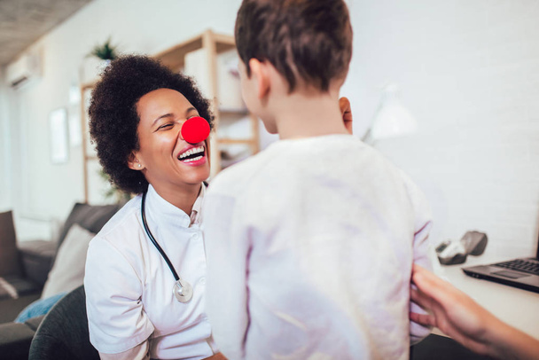 Pédiatre afro-américaine avec stéthoscope et nez de clown parlant au petit garçon  - Photo, image