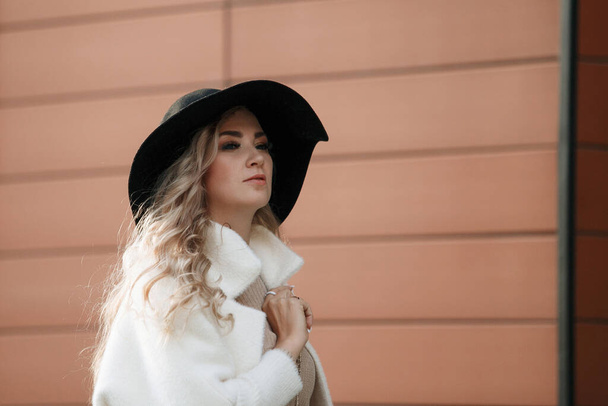 Portrait of a blonde lady in a black hat outdoors, long hair, beige coat. - Photo, Image