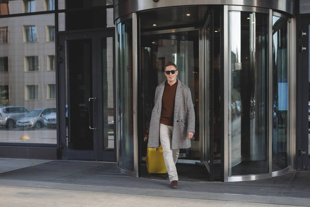 Homme en lunettes noires, avec une valise jaune en pantalon beige, vêtu d'un manteau gris, quitte l'hôtel
. - Photo, image