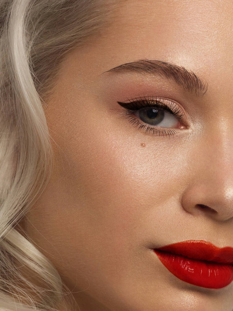 Half close-up portrait of a woman with shiny clean skin and curly blond hair. Eyeliner on the eyes, evening makeup and red lipstick. soft care, full lips, long eyelashes and thick eyebrows - Photo, image
