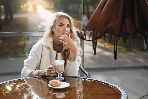 Πορτρέτο μιας ξανθιάς κυρίας σε ένα τραπέζι σε ένα καφέ τρώγοντας ένα κέικ, μακριά μαλλιά, ένα μπεζ παλτό. - Φωτογραφία, εικόνα