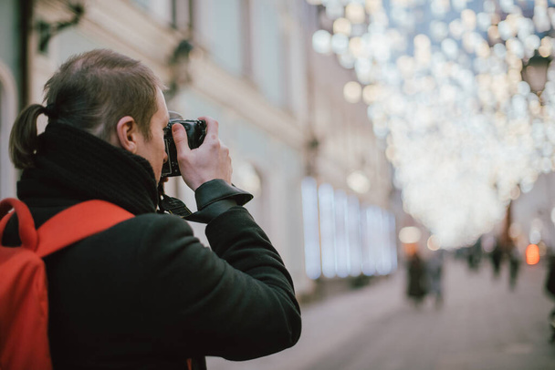 Un uomo con una macchina fotografica fotografa la strada della città vecchia in inverno. Fotografo con una macchina fotografica in cappotto e sciarpa sullo sfondo di lampioni decorati ad arte - Foto, immagini