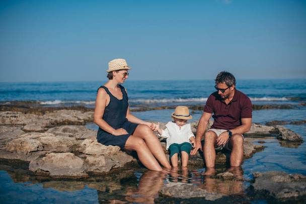 Familie mit Kind sitzen auf den Steinen vor dem Hintergrund des Meeres. junge Mutter und Vater mit einem Baby in der Nähe des Wassers vor dem Hintergrund der Stadt und das Meer am Horizont - Foto, Bild