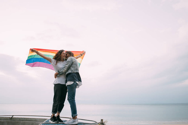 Két nő szivárvány zászlóval a parton az éggel szemben. Pár fiatal leszbikus lány szivárvány zászlót tart és boldogan ölelkezik. Egy pár leszbikus nő. - Fotó, kép