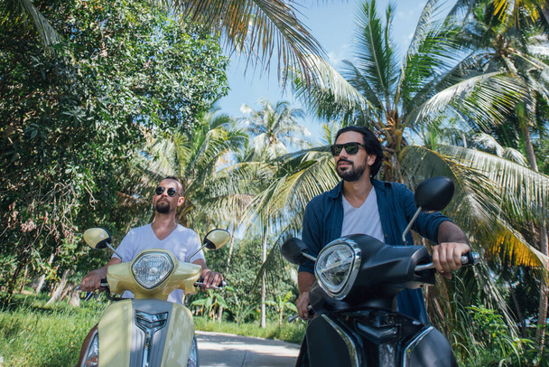 Two men on motorcycles in palm trees on a tropical island. Guys tourists on bikes on the road in a palm grove - Photo, image