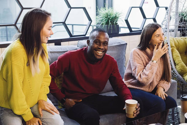 Skupina tří přátel nadnárodních mužů a žen komunikuje, usmívá se a směje se u stolu v kavárně nebo restauraci. - Fotografie, Obrázek