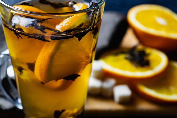 Citrus drink, tea with petals in a transparent glass mug with slices of sweet orange and refined sugar. Slicing fruit on a wooden Board with a brown napkin in a cozy dark background in a low key - Foto, imagen