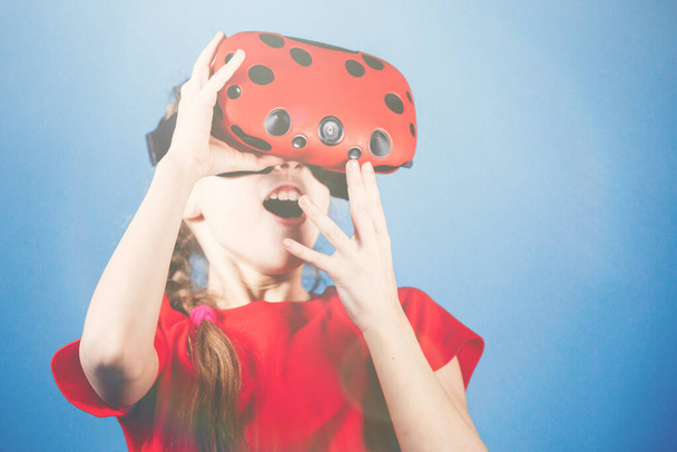 Blondje tiener meisje speelt met virtual reality bril. Computerspelletjes, blauwe achtergrond, emoties. - Foto, afbeelding