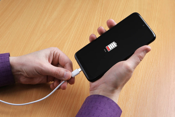 O homem conecta um cabo de carregamento a um smartphone. Bateria fraca visível no dispositivo
 - Foto, Imagem