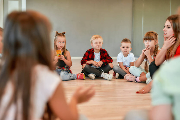Communicating with children. Full length portrait of cute little boys and girls sitting on the floor gathered around their female dance teacher and listening her carefully. Dance school - Foto, afbeelding