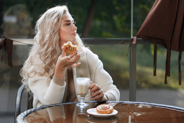 ケーキ、長い髪、ベージュのコートを食べるカフェのテーブルでブロンドの女性の肖像画。カメラのポーズをとって. - 写真・画像