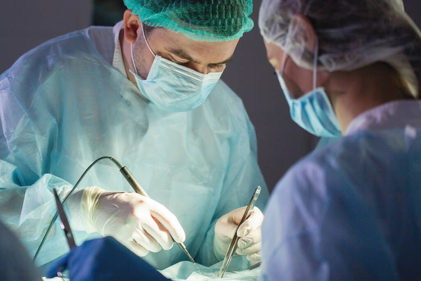 Τρεις χειρουργοί κατά τη χειρουργική επέμβαση στην κοιλιά. Μπλε στολές, γάντια λάτεξ, ιατρικά εργαλεία. Φωτισμός σημείου. - Φωτογραφία, εικόνα