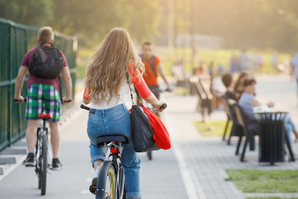 Femme fait du vélo sur une piste dans un parc de la ville. Cheveux longs, jeans, chemise rouge et blanche. - Photo, image