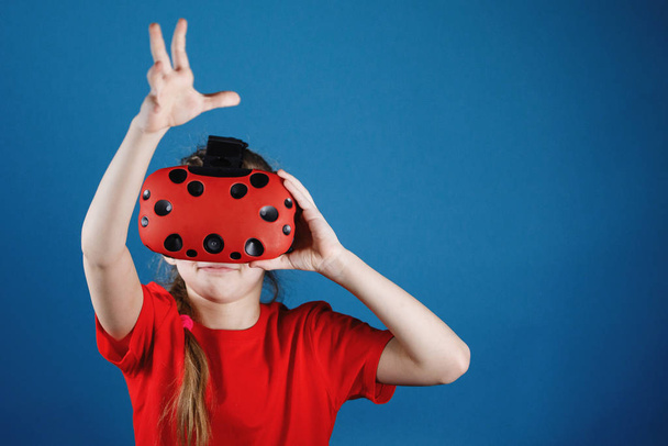 Blondy teen girl plays with virtual reality glasses. Computer games, blue background, emotions. - Fotó, kép