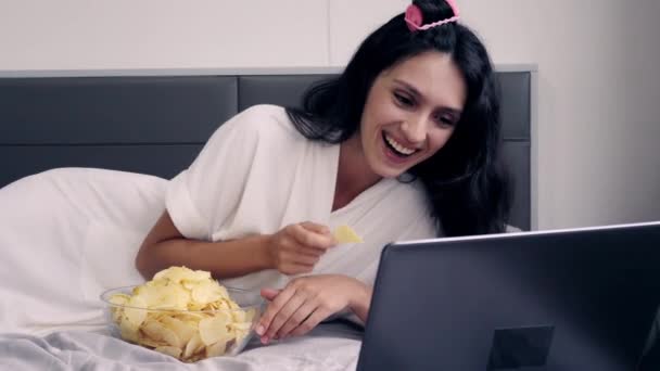 Beautiful woman laughing so much when she watch comedy movie and eat snack or potato chips on bed on holiday weekend by using notebook computer. She wear cloak, get hair curler. Pretty girl get happy - Materiaali, video