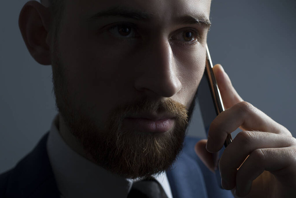 Close-up, studio dramatic portrait of a handsome bearded man, in a business suit, holding a new smartphone in his hand, against a tuxedo background. On a gray background. Office Style. Business design and style. New fashionable smartphone - Fotó, kép