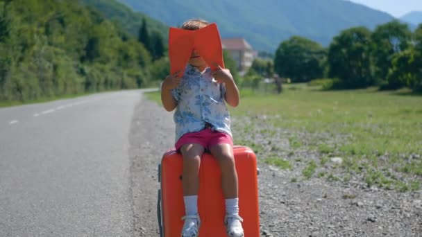 Cute toddler boy with suitcase and flippers is having fun waiting for the journey and waving his flippers in the fresh air along the road. Leisure, weekend, travel and tourism concept - Footage, Video