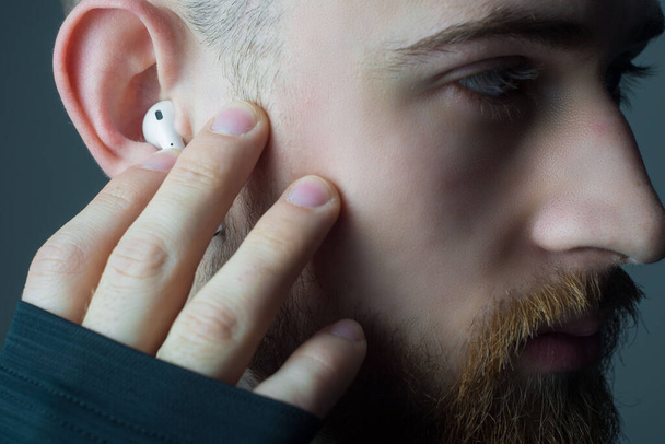 Kulağında kablosuz beyaz kulaklıkla kameraya bakan 25 yaşındaki genç sakallı bir adamın yakın plan portresi. Kulaklıkların reklam fotoğrafı. Kulaklıklı bir sporcu. El ele tutuşup müzik dinliyor. - Fotoğraf, Görsel