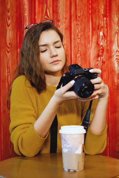 One young pretty woman in glasses, with long hair in a mustard sweatshirt with a camera, sits in a cafe against the background of a red wooden wall, photographs a white glass on the table. Lifestyle, casual, joy of communication, student life. - Photo, Image