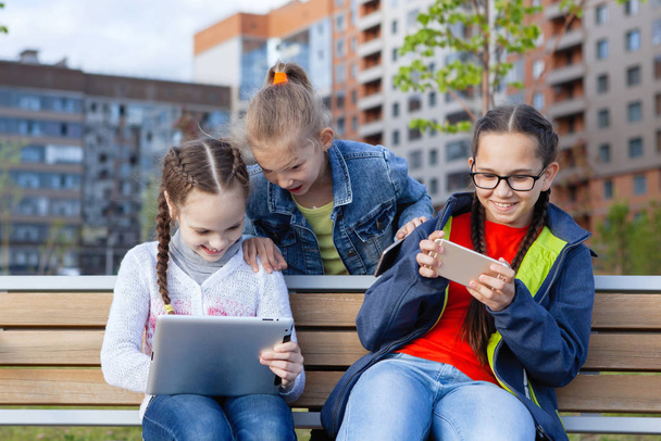Tři dospívající dívka s chytrým telefonem sedí na lavičce v letním městském parku. - Fotografie, Obrázek