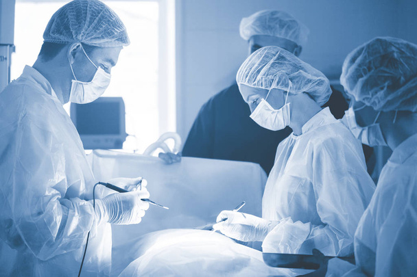 Три хирурга во время операции на животе. Синяя униформа, латексные перчатки, медицинские инструменты. Точечное освещение. - Фото, изображение