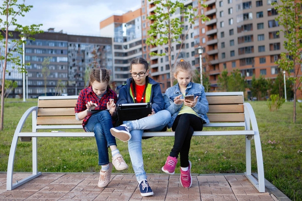 Tre adolescenti con smartphone e tablet seduti su una panchina in un parco cittadino in estate
. - Foto, immagini