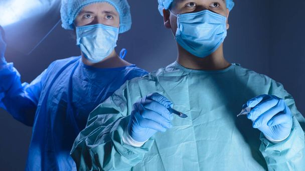 Portret dwóch chirurgów w sterylnych fartuchach, kapeluszach i maskach. Jeden chirurg, przygotowujący się do operacji, trzyma skalpel z uchwytem na igłę w rękach, inny lekarz trzyma prześwietlenie pacjenta, kierując jego oczy prosto. W związku z tym  - Zdjęcie, obraz