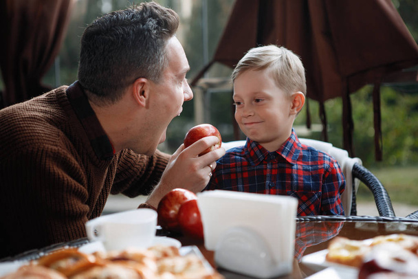 Isä ja poika istuvat kahvilan pöydässä. Mies tarjoaa pojalle omenaa. Vanhempien rakkaus, onnellinen lapsuus, lastenhoito
. - Valokuva, kuva