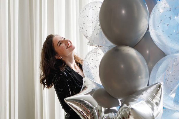 Junge hübsche Frau mit Luftballons drinnen vor dem Hintergrund eines großen Fensters mit Vorhängen. Bälle sind blau, weiß und golden. Festliche Überraschung, Geburtstagsgeschenk. - Foto, Bild