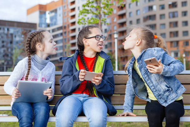Три девочки-подростка со смартфоном и планшетом сидят на скамейке в городском парке летом
. - Фото, изображение