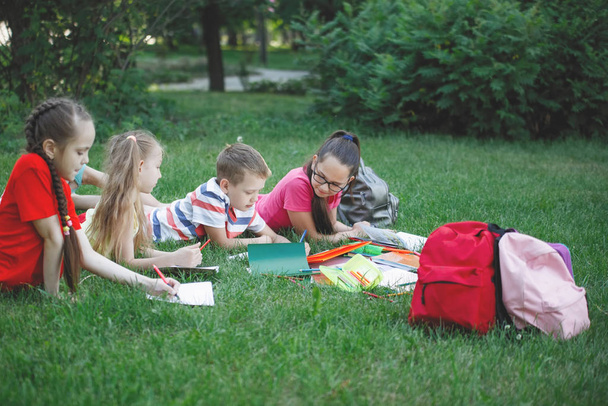 Τέσσερα παιδιά κείτονται στο γρασίδι στο πάρκο της πόλης. Διαβάζουν βιβλία και ζωγραφίζουν σε άλμπουμ. - Φωτογραφία, εικόνα