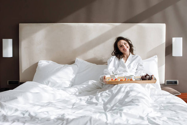 Yatağında yemek tepsisi olan genç bir kadın sabah uyandığında gülümsüyor ve hayatın tadını çıkarıyor.. - Fotoğraf, Görsel