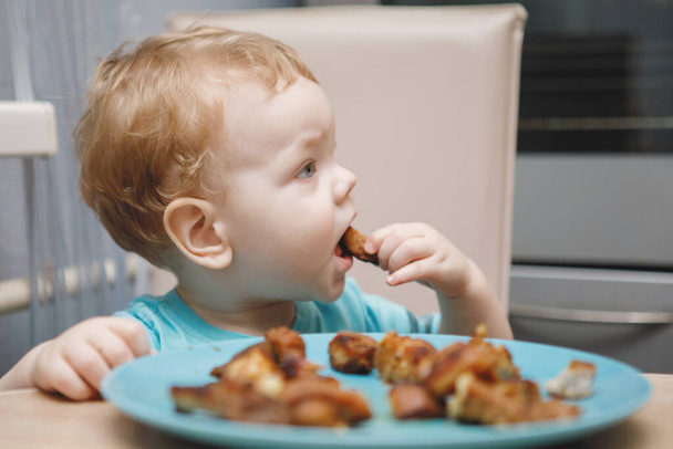 Egy kisbaba szőke fiú sült húst eszik egy tányérról az asztalnál.. - Fotó, kép