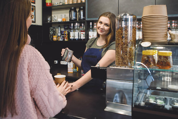 Une Barista dans un tablier sert du café dans une tasse de carton à un client au comptoir du bar.. - Photo, image