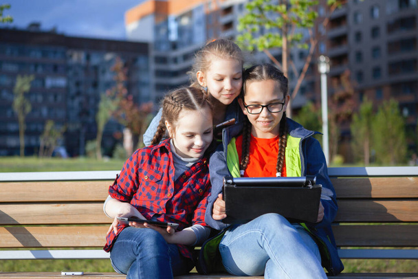 Tre adolescenti con smartphone e tablet seduti su una panchina in un parco cittadino in estate
. - Foto, immagini