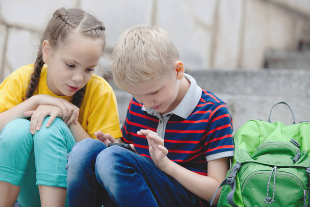 Two schoolchildren: a girl with pigtails and a blond boy look at a smartphone, sit on the steps on a sunny summer day outdoors. - Fotó, kép