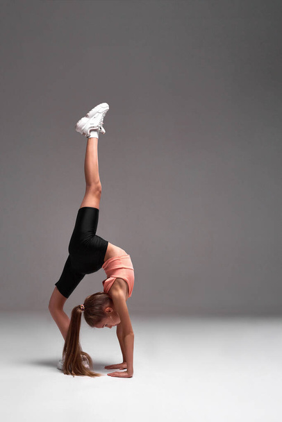 Get active. Flexible cute little girl child bending over backwards while stretching her body isolated on a grey background. Sport, training, fitness, active lifestyle concept. Vertical shot. - Фото, изображение