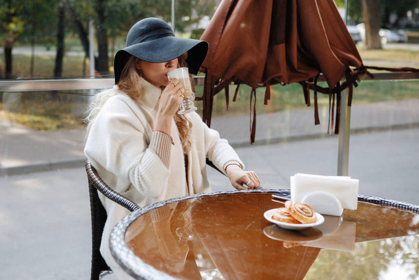 Portrét blondýny v černém klobouku u stolu v kavárně popíjející latté, dlouhé vlasy, béžový kabát. - Fotografie, Obrázek