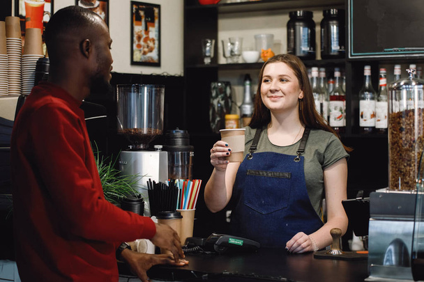 Afrikaanse man koopt een kop koffie bij een bar balie bij een barista in een cafe. - Foto, afbeelding