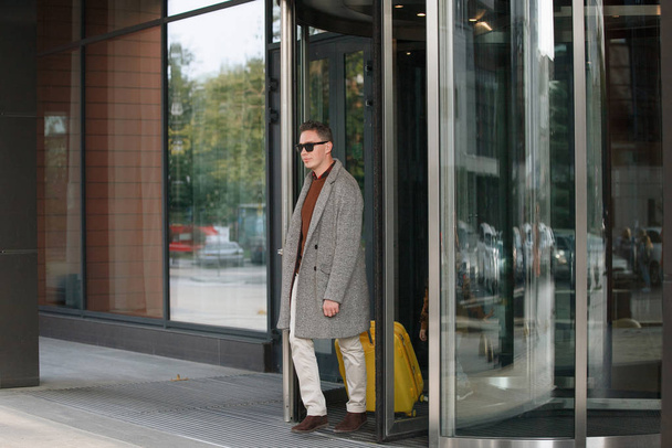 Man in black glasses, with a yellow suitcase in beige trousers, in a gray coat, leaves the hotel. - Fotó, kép