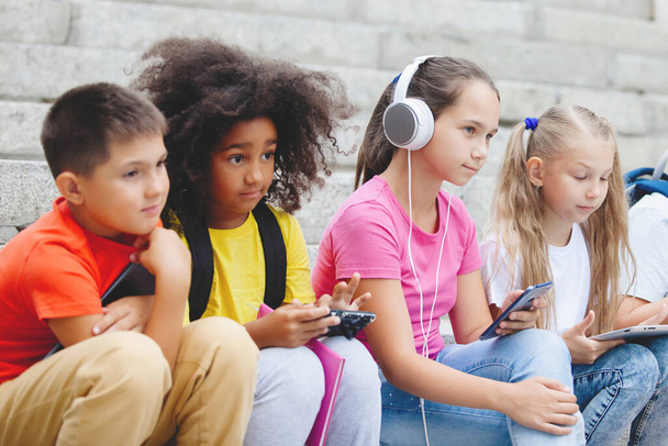 Grupo de escolares de diferentes nacionalidades, em roupas coloridas sentado em degraus de pedra. Adolescentes conversando, ouvindo música em fones de ouvido, lendo livros
. - Foto, Imagem