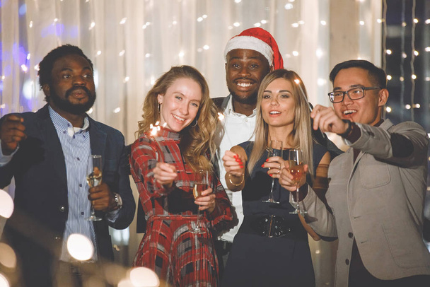 Skupina multietnických lidí stojí s sklenicemi šampaňského a jiskry na svátcích v čepicích a smějí se. Vybrané, měkké zaměření. Vintage efekt starého filmu. Filtr pro hluk a zrní. - Fotografie, Obrázek