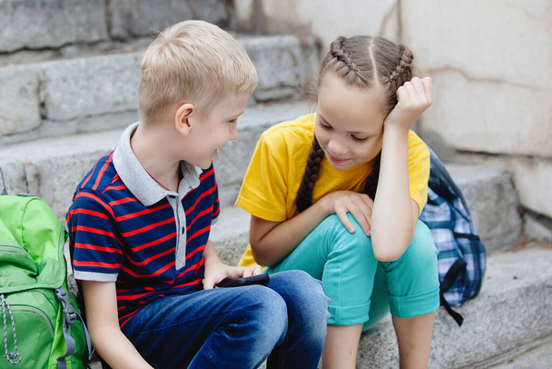 Δύο μαθητές: ένα κορίτσι με κοτσίδες και ένα ξανθό αγόρι κοιτάζουν ένα smartphone, κάθονται στα σκαλοπάτια σε μια ηλιόλουστη καλοκαιρινή μέρα στο ύπαιθρο. - Φωτογραφία, εικόνα