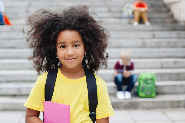 Портрет африканської дівчини - підлітка з кучерявим чудовим волоссям. Шкільна дівчина У жовтій футболці з рожевим записником, з рюкзаком. Вибіркове зосередження. - Фото, зображення